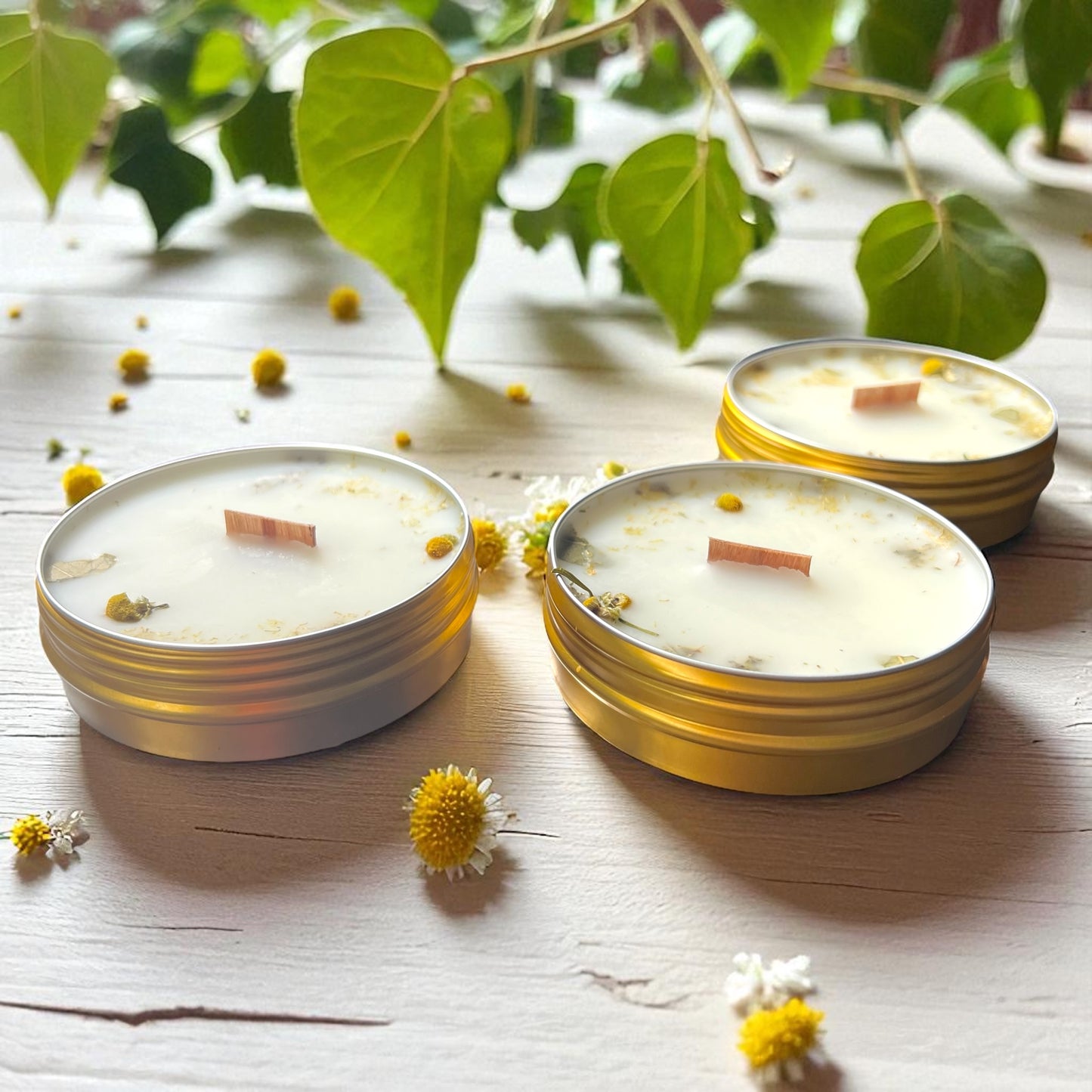 Three small candles in gold tins on a wooden surface with flowers and leaves.