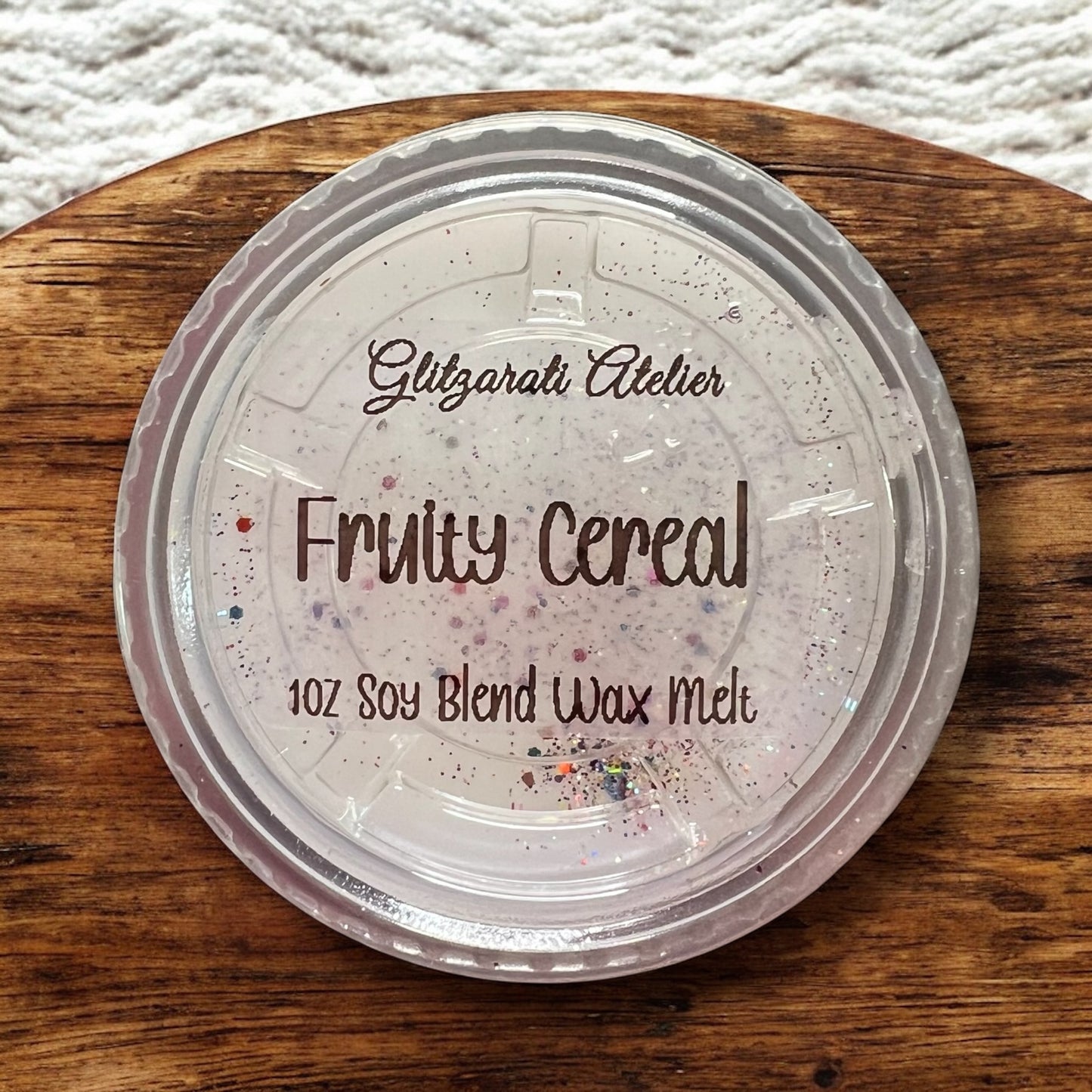 Wax melt container labeled 'Fruity Cereal' by Glitzerati Atelier on a wooden surface.