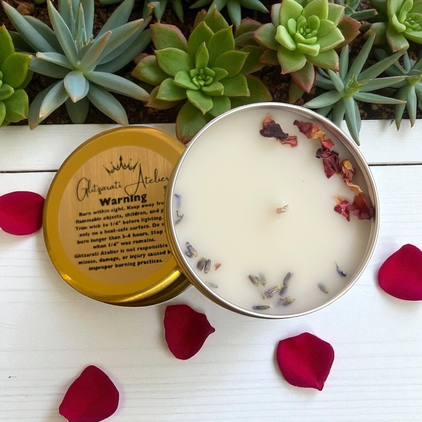 Candle in a tin with dried flowers surrounded by succulents and red petals on a white surface