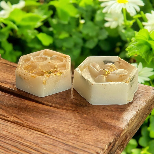 Two hexagonal honeycomb-shaped soap bars on a wooden surface with a natural background of greenery and flowers.