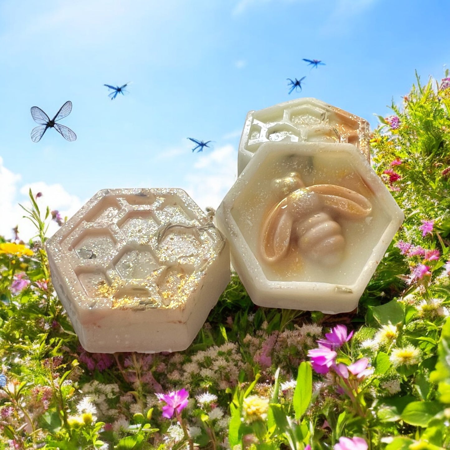 Three hexagonal honeycomb-shaped soaps with bee designs on a grassy background with flowers and dragonflies.