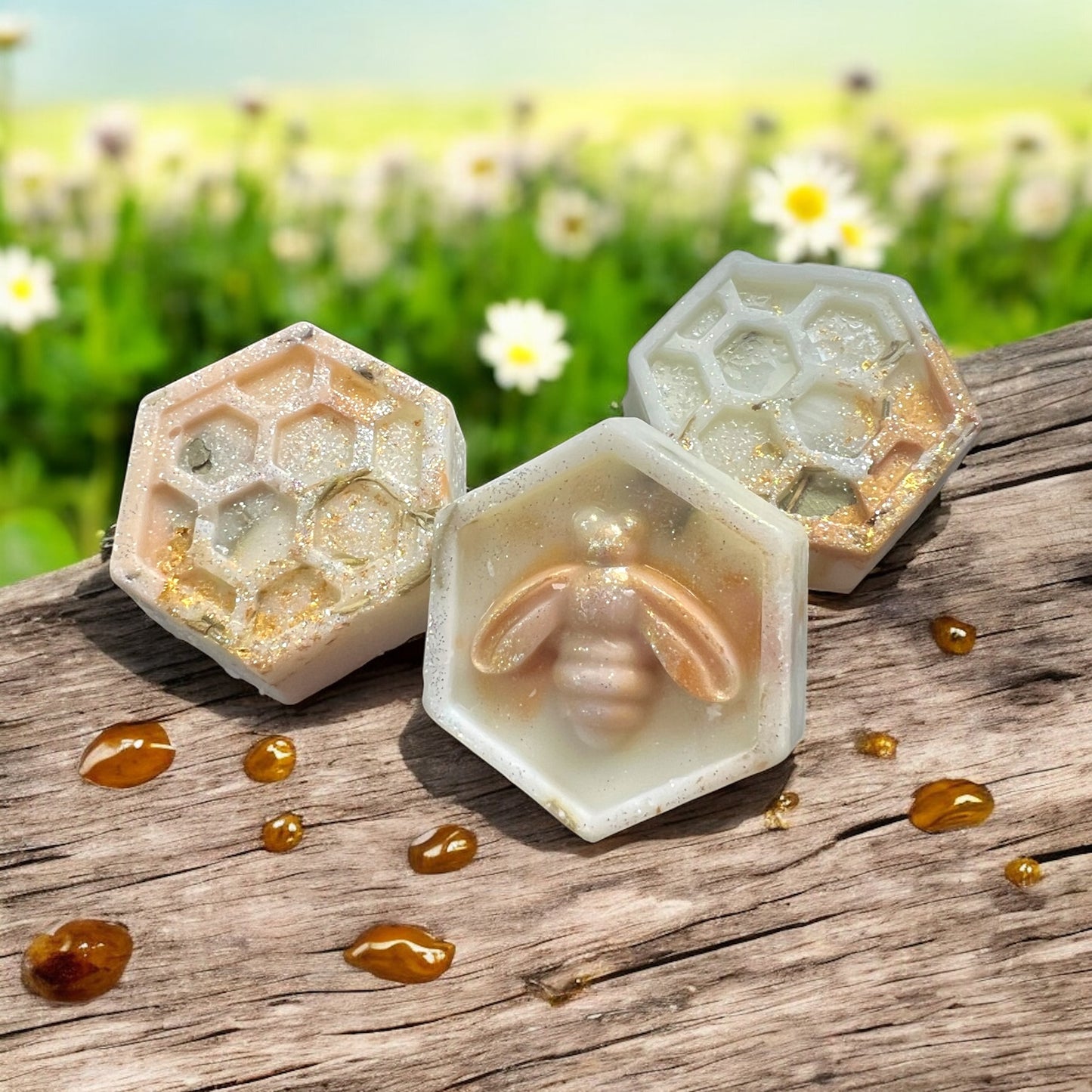 Three honeycomb-shaped soap bars with a bee design on a wooden surface with a field of flowers in the background.