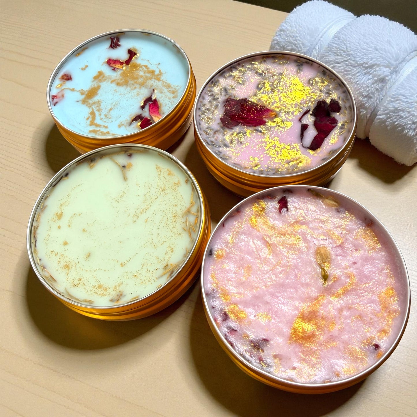 Four small containers of bath salts with decorative elements on a wooden surface, accompanied by white towels.