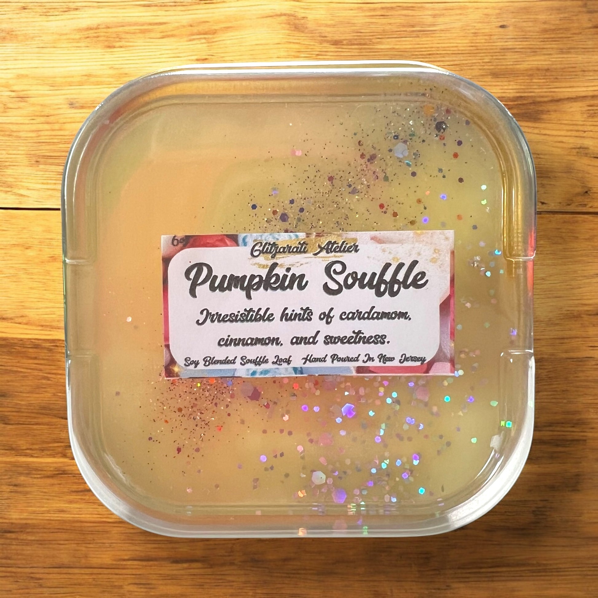 A collection of Glitzarati Atelier's scented wickless wax melt souffles in various colors, presented in aluminized tins surrounded by colorful flowers.