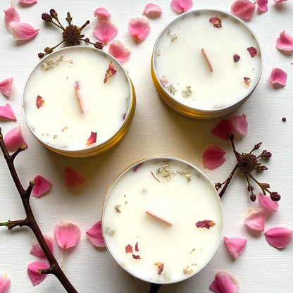 Three white candles with floral elements on a white surface with pink petals and branches.