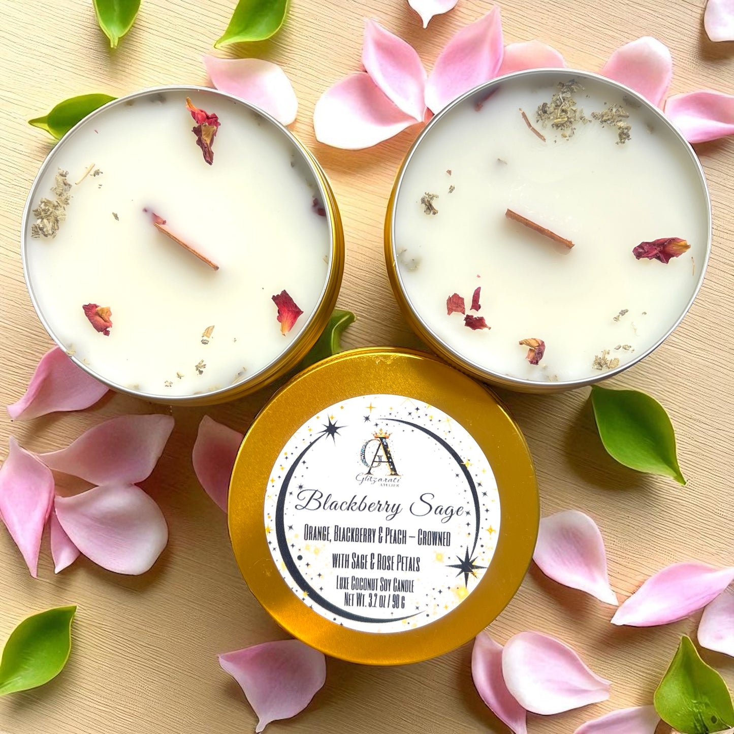 Two candles in gold tins with floral petals and leaves on a wooden surface.