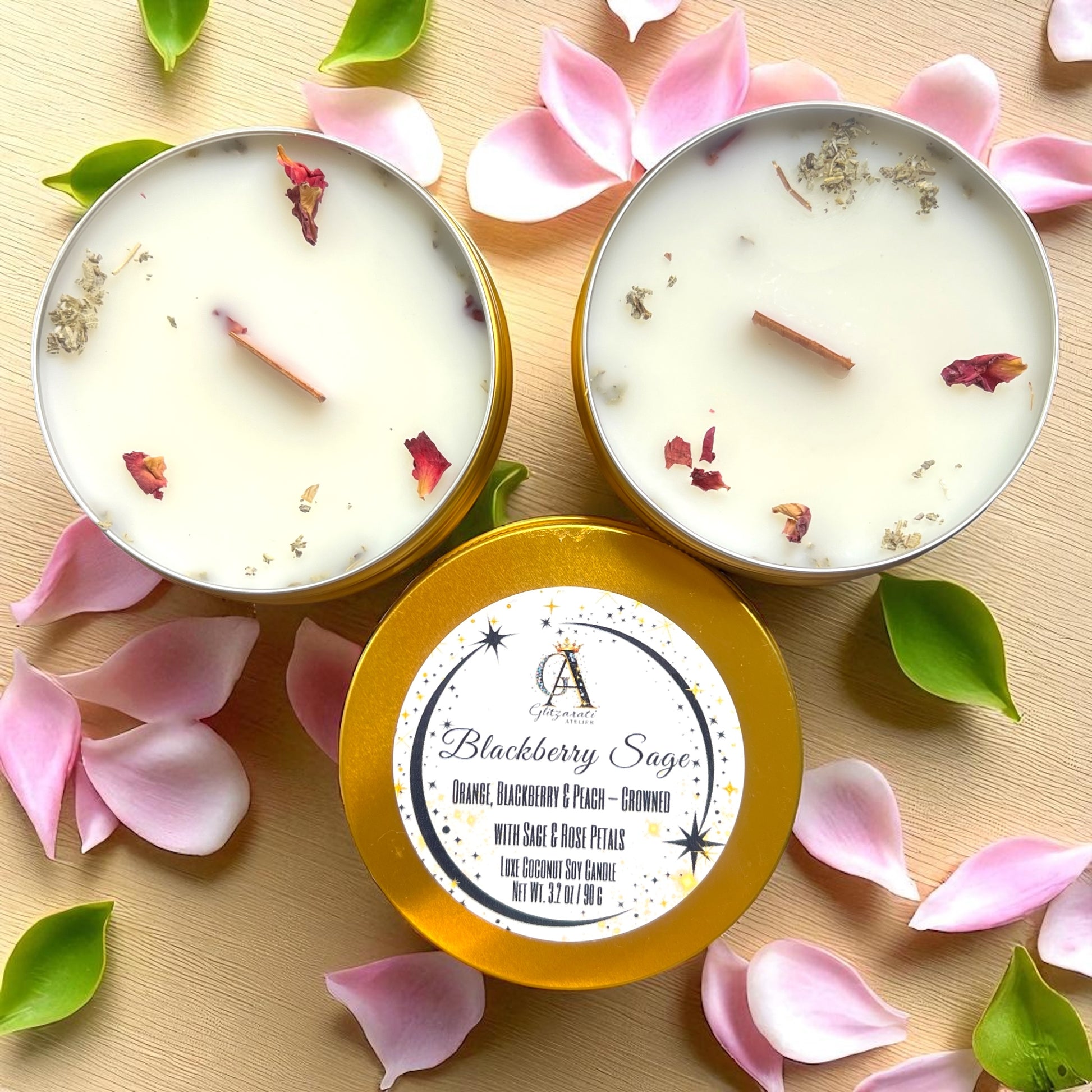 Two candles in gold tins with floral petals and leaves on a wooden surface.