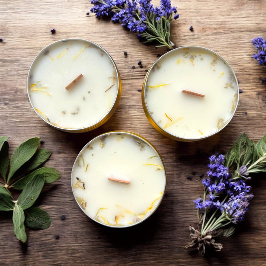 Three small candles in tin containers on a wooden surface with lavender and sage leaves.