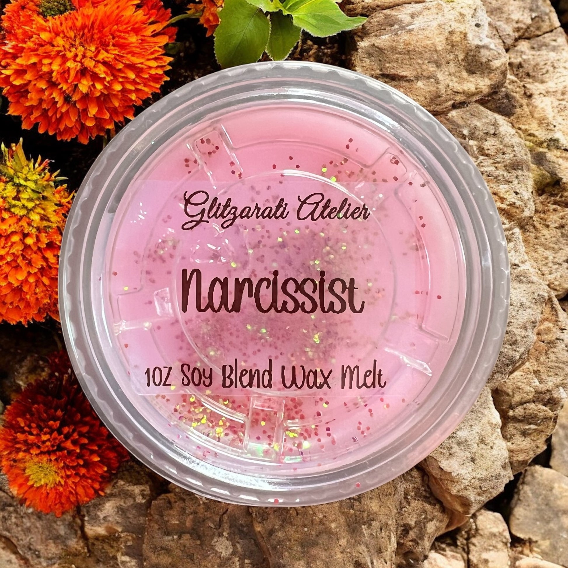 Pink wax melt container labeled 'Narcissist' by Glitzerati Atelier on a rocky surface with orange flowers.