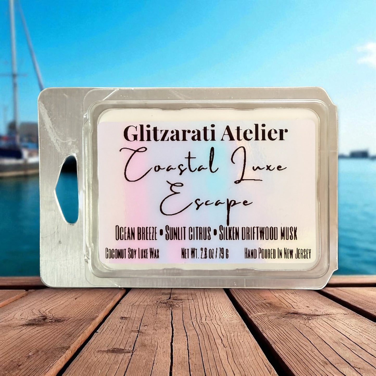 Wax melt package with 'Glitzerati Atelier Coastal Luxe Escape' label on a wooden surface with water background