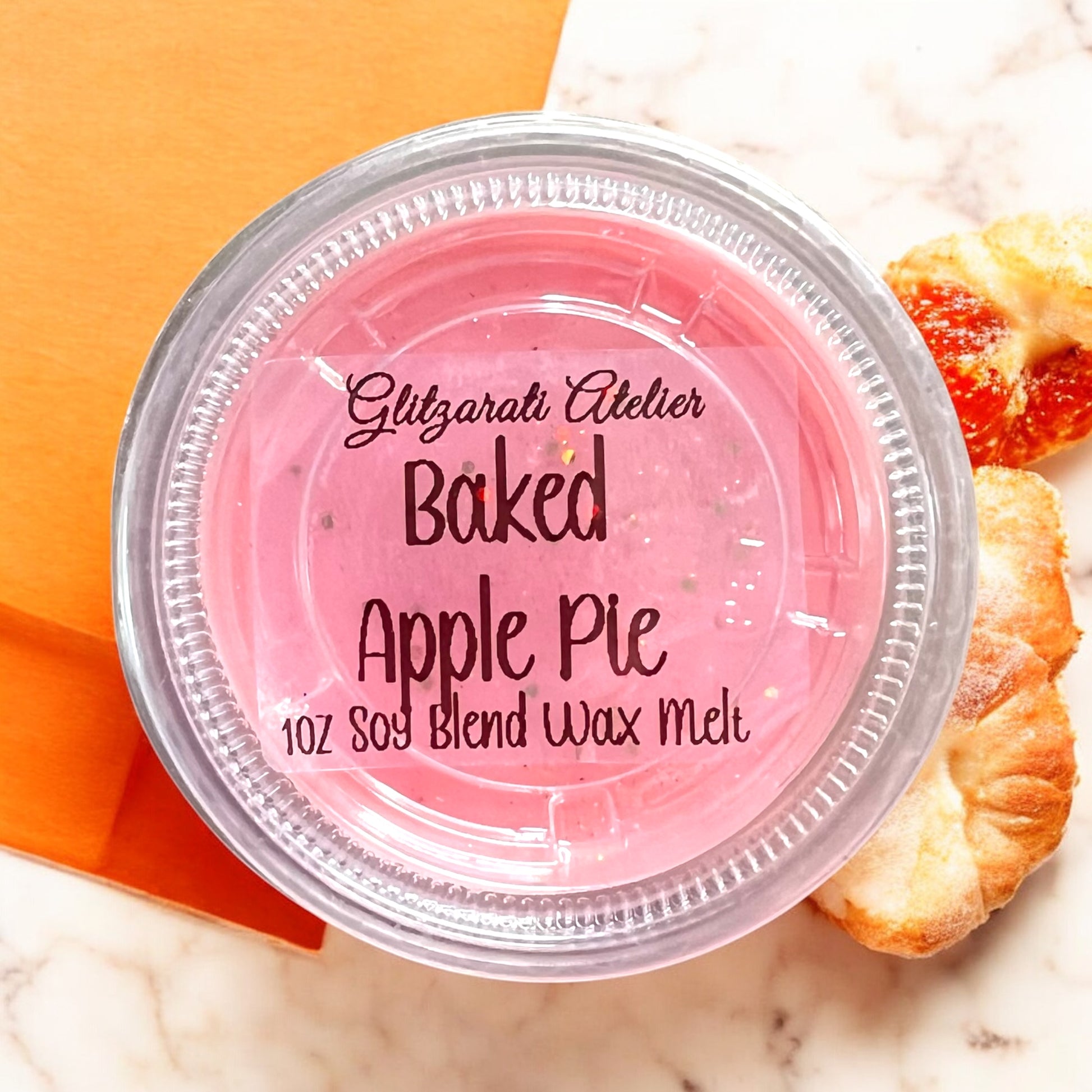 Pink wax melt container labeled 'Baked Apple Pie' by Glitzerati Atelier on a marble surface with a decorative apple pie.