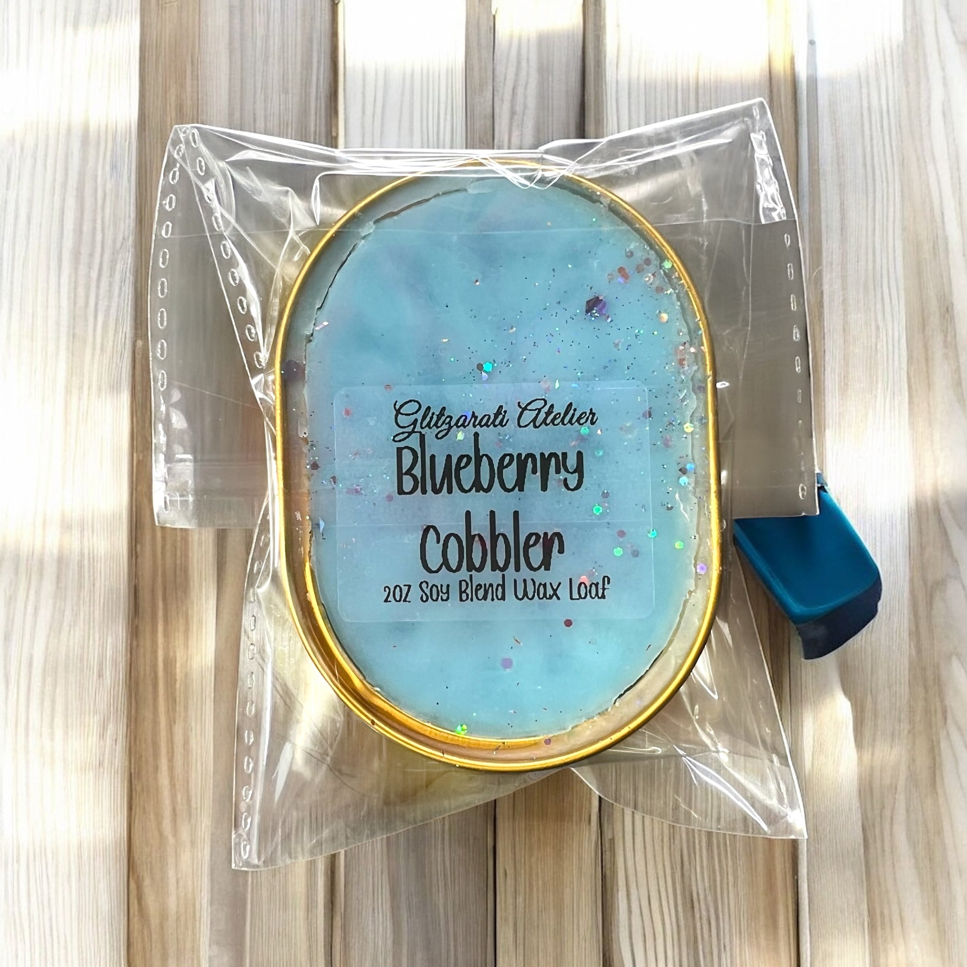 Wax melt container labeled 'Blueberry Cobbler' by Glitzyari Atelier in clear packaging on a wooden surface.
