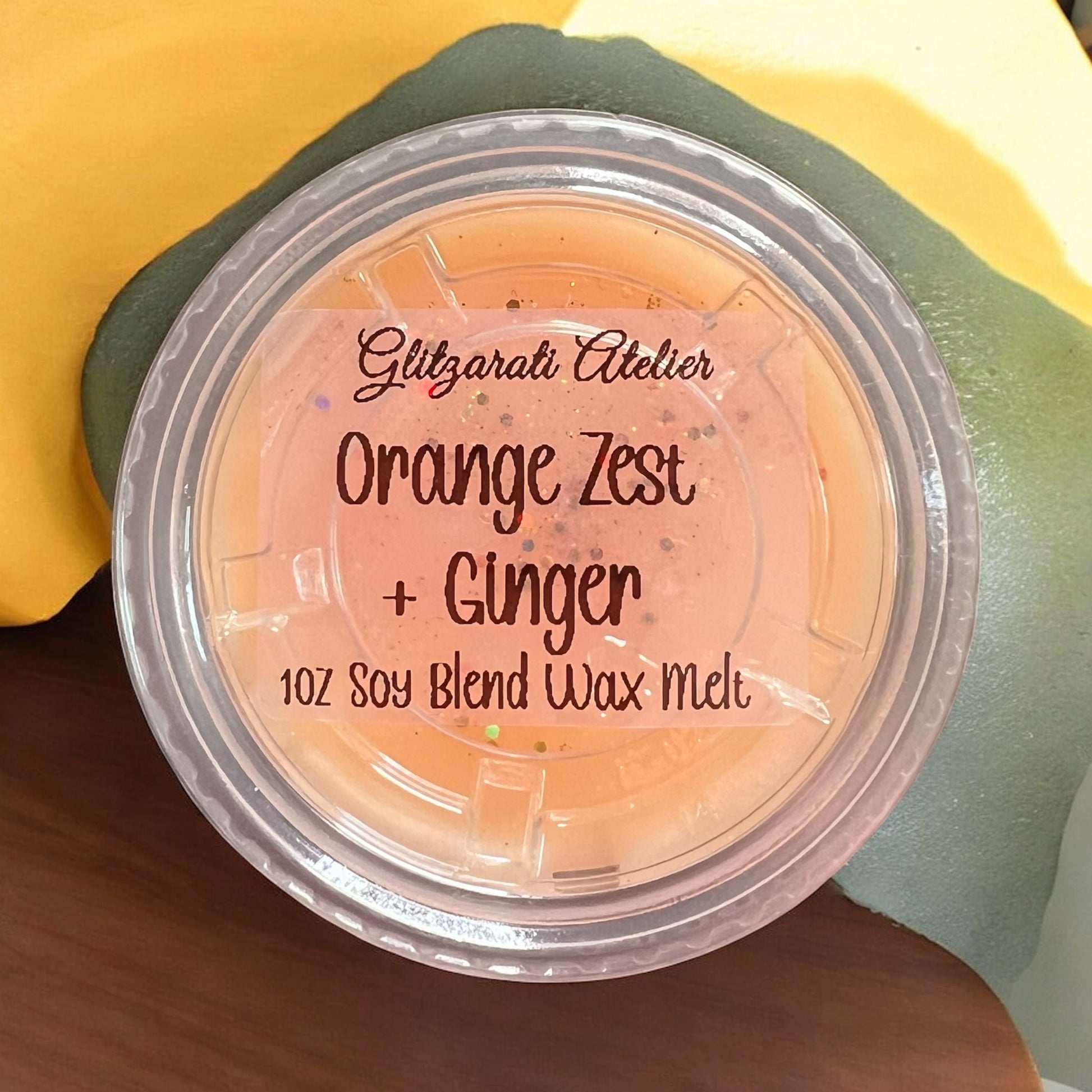 Wax melt container labeled 'Orange Zest + Ginger' by Glitzarati Atelier on a hand.