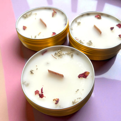 Three small candles in gold tins with dried flowers on a pink and purple gradient background