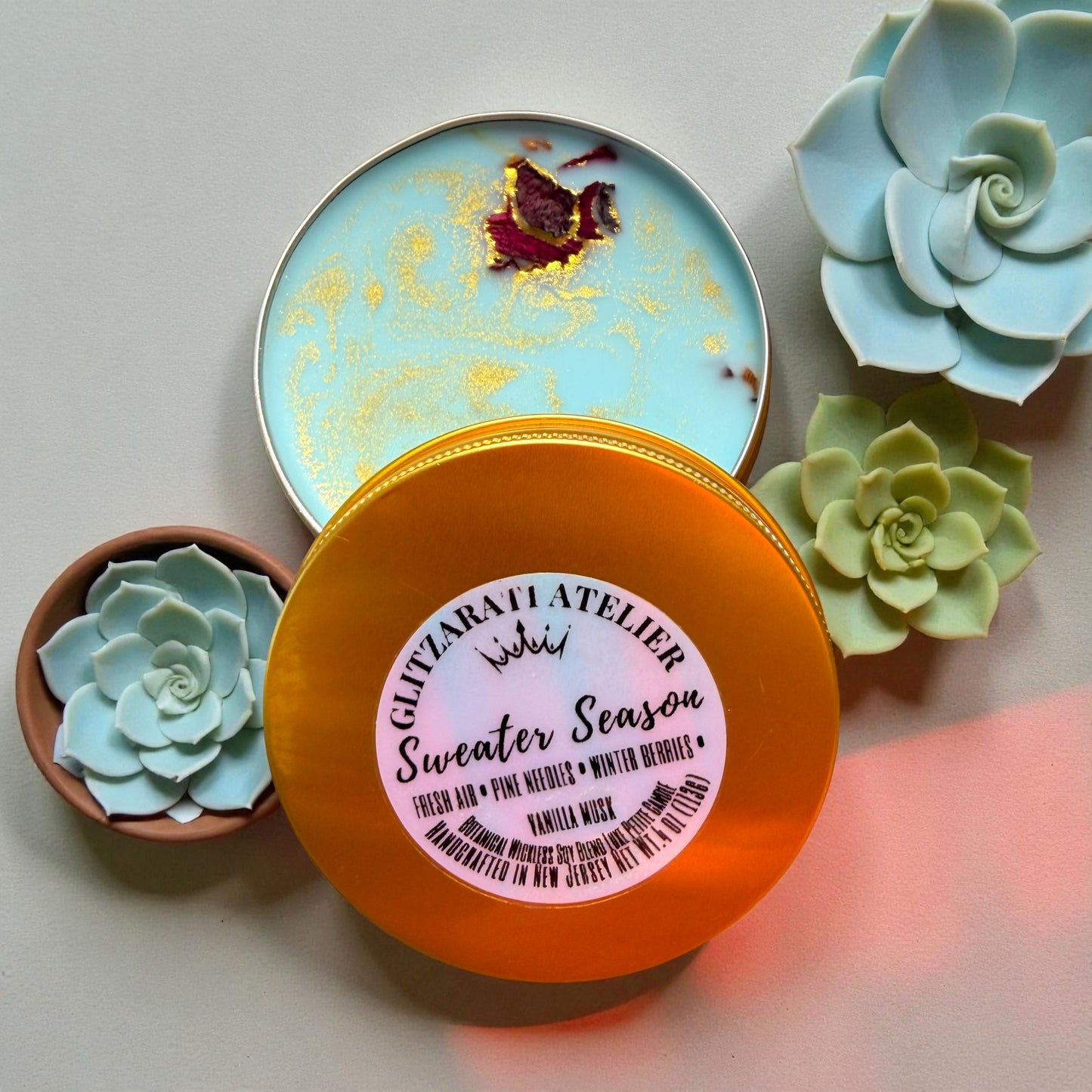 Orange container with a label surrounded by small succulent plants on a light background