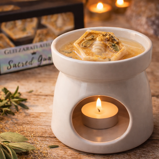 White ceramic oil burner with a lit candle on a wooden surface, surrounded by herbs and a box labeled 'Sacred Incense'.