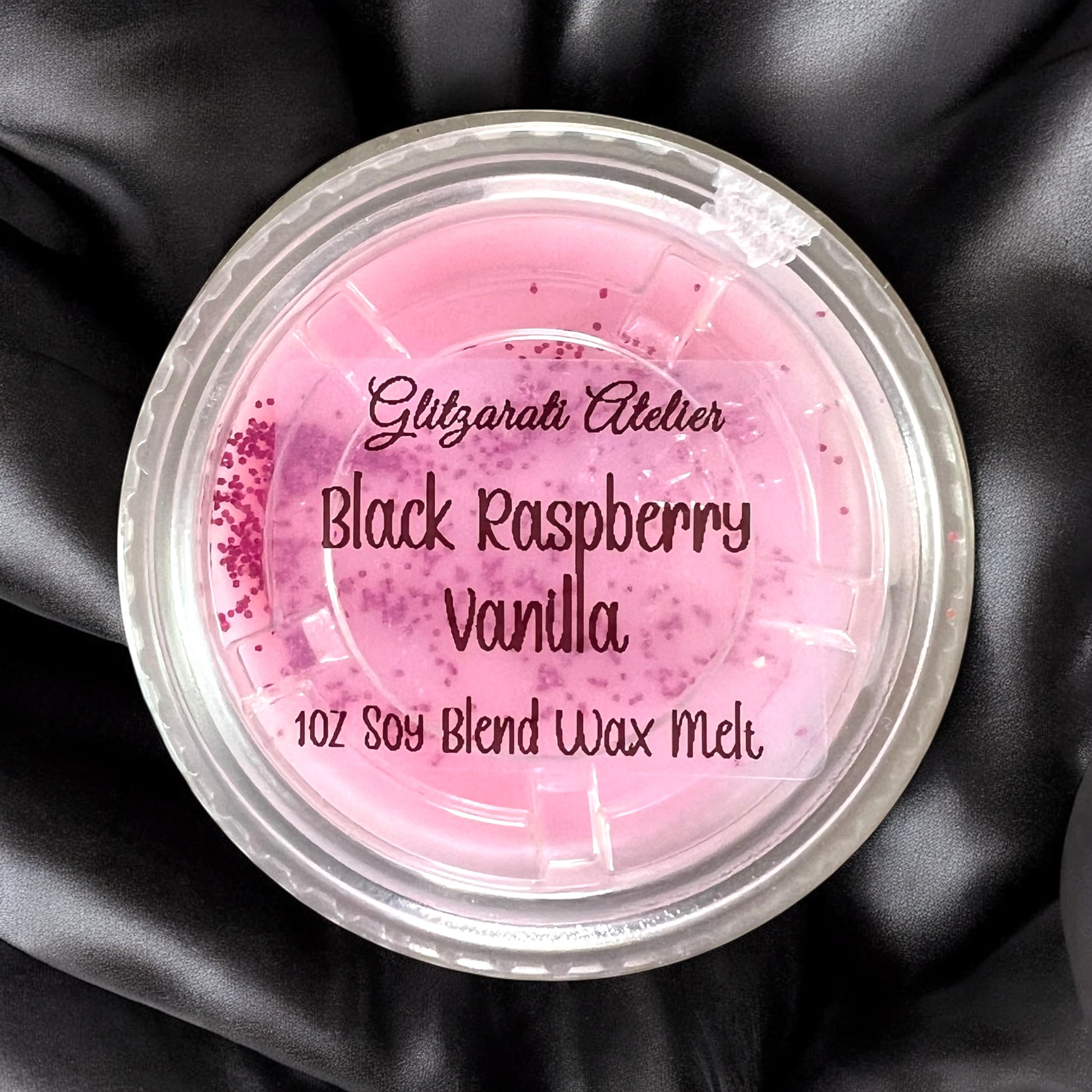 Pink wax melt container labeled 'Black Raspberry Vanilla' on a black satin background