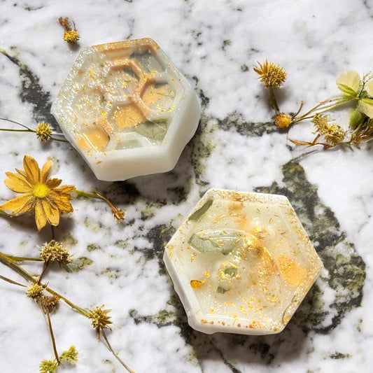 Two hexagonal soaps with natural elements on a marble surface with flowers.