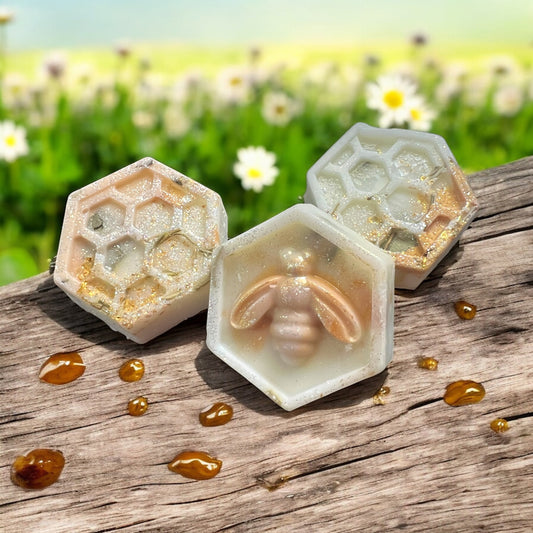 Three honeycomb-shaped soap bars with a bee design on a wooden surface with a field of flowers in the background.