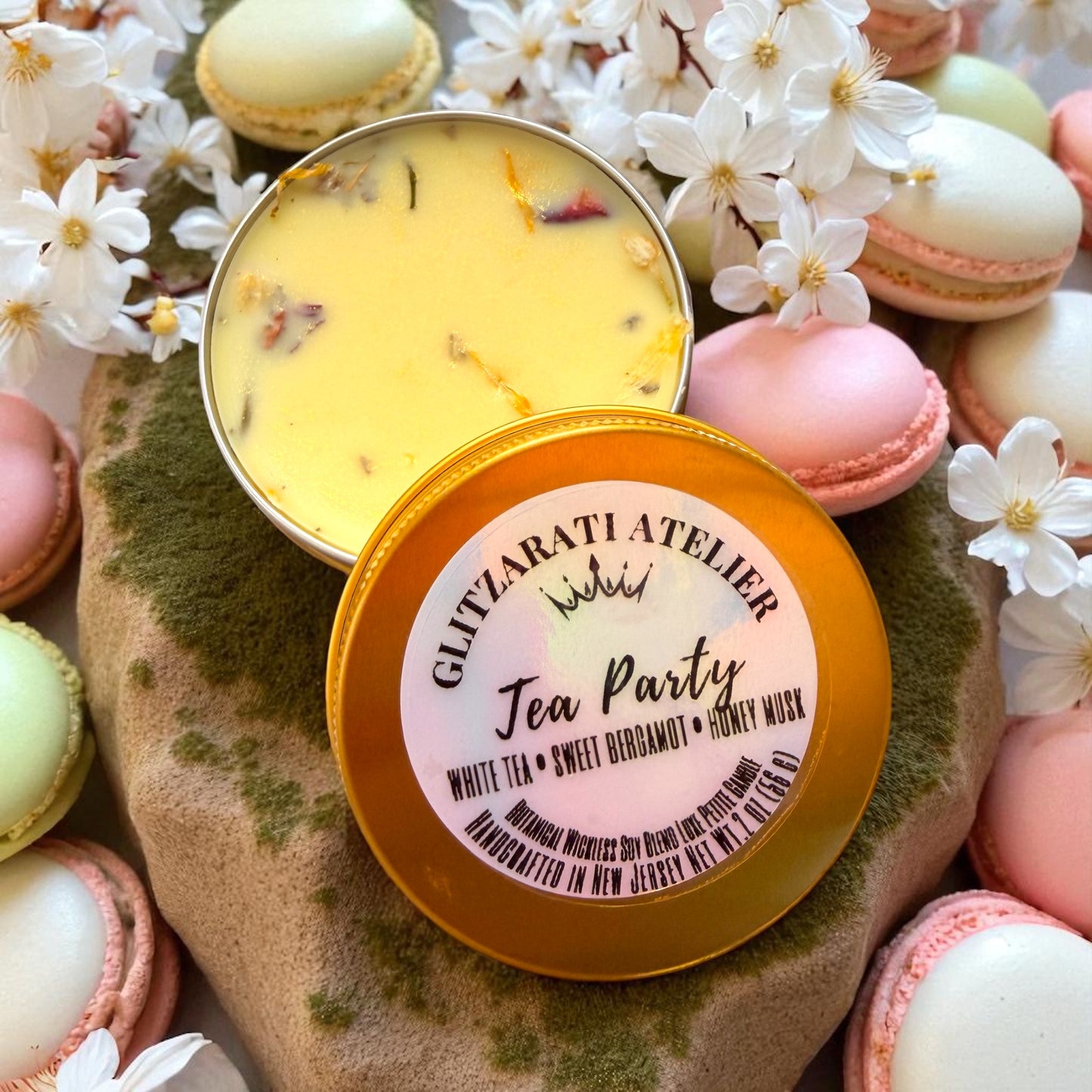 Two tins of 'Tea Party' scented candles with macarons and flowers on a stone surface.