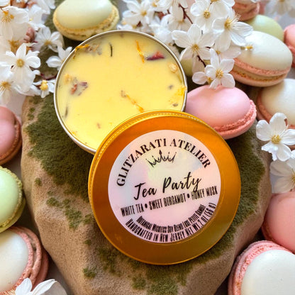 Two tins of 'Tea Party' scented candles with macarons and flowers on a stone surface.