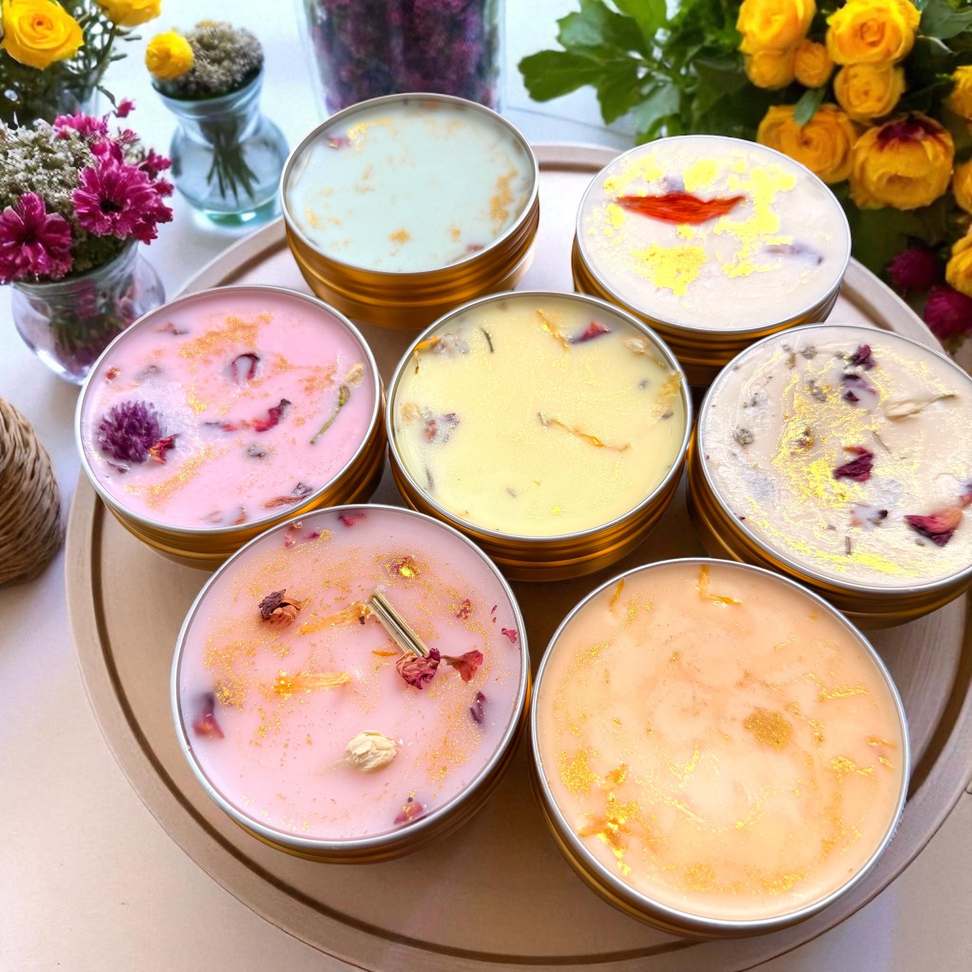 Assorted candles in tins with floral decorations on a round tray, surrounded by flowers.