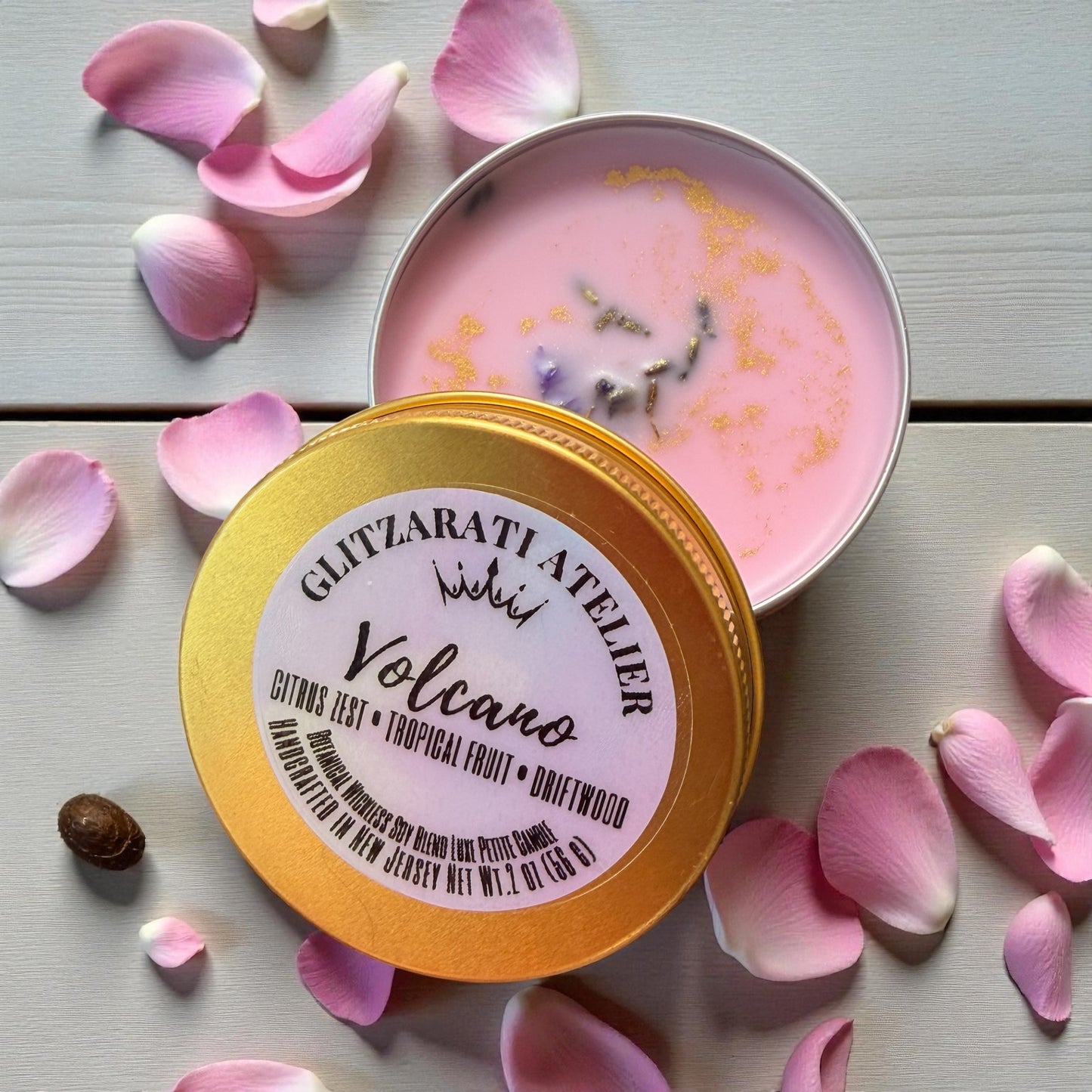 Pink candle in a tin with a gold lid surrounded by pink petals on a light wooden surface