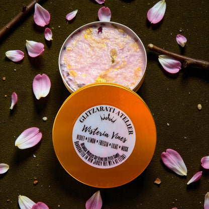 Open container of Ghyllarati Atelier Wisteria Vines with orange lid and pink interior, surrounded by pink petals on a dark background.